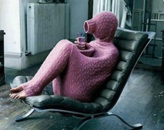 A person wearing a textured, pink, full-body hooded outfit sits in a black leather chair, holding a small object in their hands.