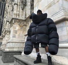 A person wearing an oversized black puffer jacket with a hood and thick gloves stands on stone steps in front of a grand, ornate stone building.