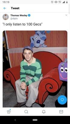 A young man wearing a green and white striped sweater and a blue cap sits on a red, curved couch with spiral armrests. Behind him, a large blue plush toy with a smiling face sits on a shelf, and a purple plush toy with big eyes sits on the right side of the couch.