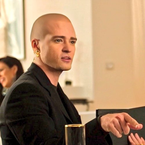 A bald man in a black suit with a gold earring looks forward, pointing with his right hand while holding a gold-rimmed glass.