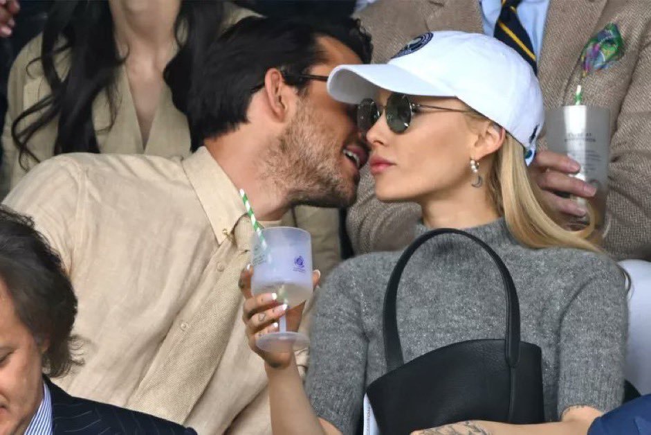 A man and a woman are seated together at an event, the man kissing the woman's cheek while she looks away, holding a drink with a straw.