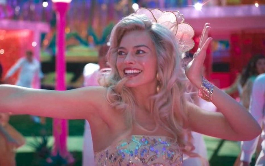 A smiling woman with blonde hair and a glittery pink and blue sequined dress waves enthusiastically at a vibrant, colorful party scene with pink lighting and dancing people in the background.