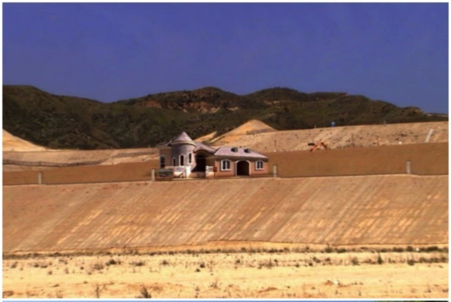A small, pinkish building with a dome sits atop a large, dry, sloped earth embankment under a clear blue sky, with green hills in the background.