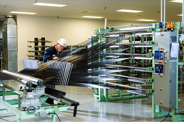 A worker in a hard hat monitors a large industrial machine weaving metal rods into a grid pattern in a brightly lit factory.