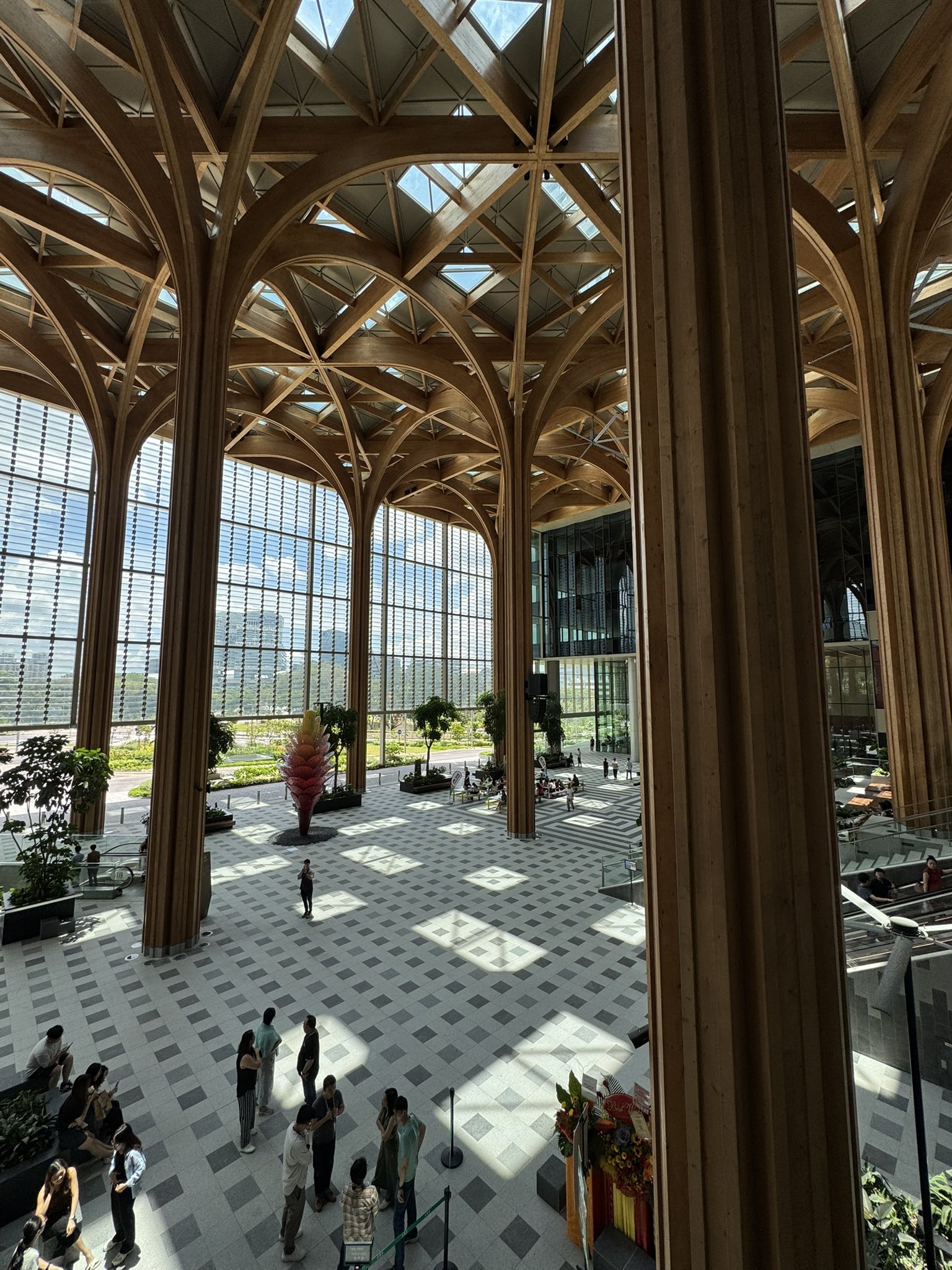 A spacious, modern atrium with a soaring wooden lattice ceiling and large arched windows, featuring a checkered floor, scattered visitors, and indoor plants, bathed in natural sunlight.