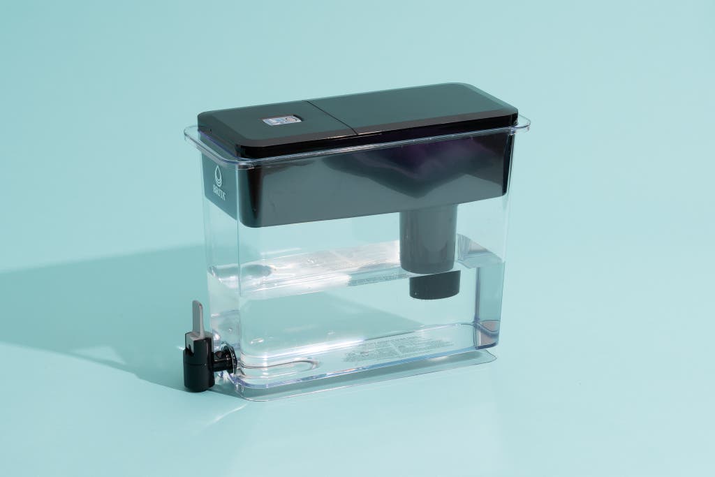 A clear plastic water filter pitcher with a black top and spigot, filled with water, sitting on a light blue surface.