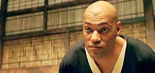 A bald man with a serious, intense expression wears a dark martial arts uniform with a white collar, set against a wooden, traditional-style background.