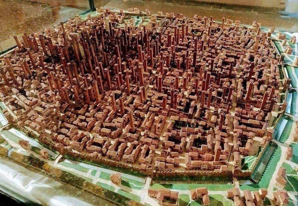 A detailed 3D model of a dense urban area, featuring hundreds of reddish-brown buildings with varying heights, surrounded by green spaces and roads, displayed under glass.