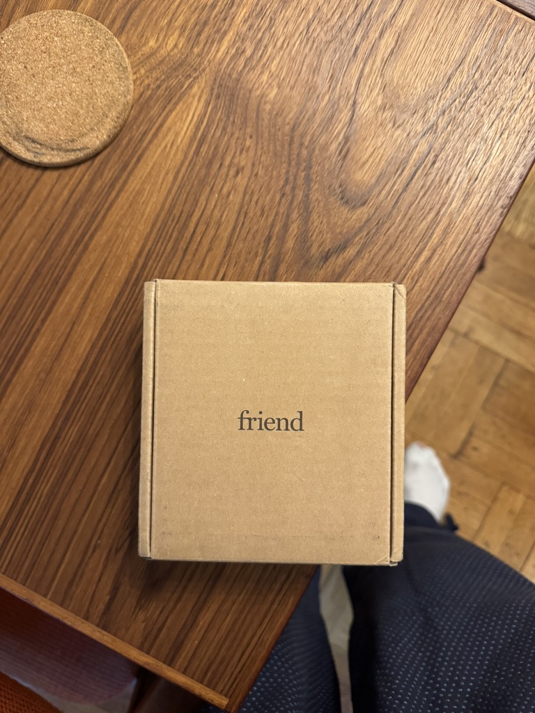 A small, square brown cardboard box with the word "friend" printed on it sits on a wooden table, next to a round cork coaster.