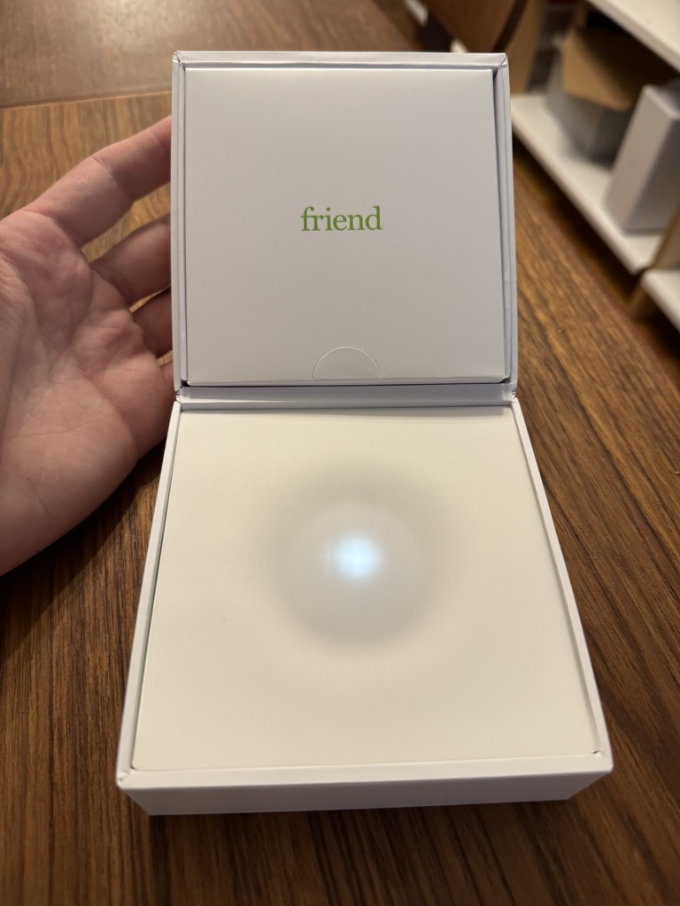 A hand holds an open white box with the word "friend" in green text on the lid, revealing a glowing light inside the box on a wooden surface.