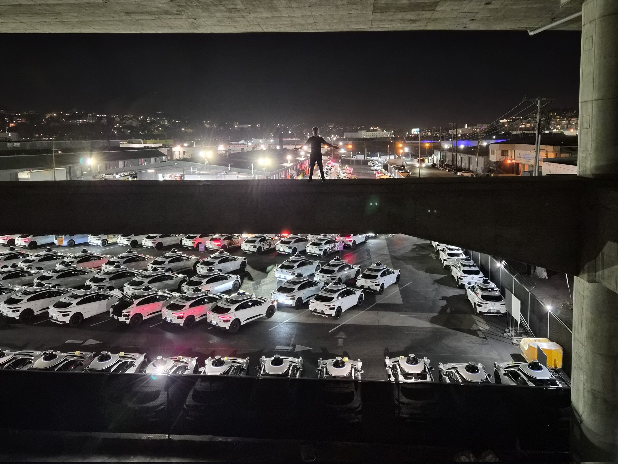 At night, a lone figure stands on a concrete overpass overlooking a large parking lot filled with numerous white self-driving taxis, with city lights visible in the background.